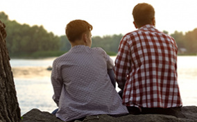 A father and son sitting on a rock face away from the camera and look at a river in front of them.