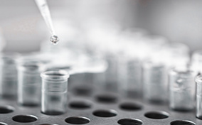 A syringe drops a liquid sample into a test tube as it sits in a rack.