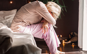 A woman sits on the edge of a bed with her feet on the ground and folds over her knees, resting her forehead on her folded arms.