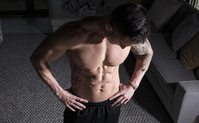 A shirtless man looks downward and holds his hands on his hips.