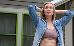A woman in a sports bra and button down stands outside a house and holds her hands behind her head.
