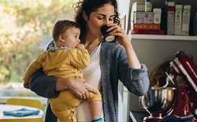 A mother holds a baby in her right arm while drinking out of a cup in her left hand.
