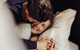 A couple cuddle in bed while smiling at each other.