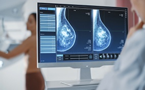 A person in the background is in a mammogram machine while a doctor in the foreground views the results on a screen.
