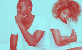 A couple sits on the edge of a bed facing away from each other and looking concerned.