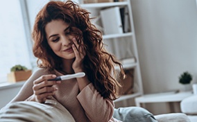A woman sits on a sofa smiling as she holds a pregnancy test.