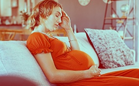 A pregnant woman sits on the sofa with her eyes closed and her hand pressed to her forehead.