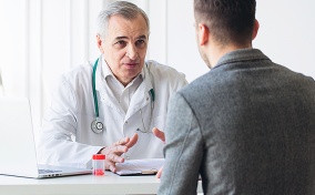 A doctor in a white coat and stethoscope gesticulates at he talks to a patient with his back to the viewer. 