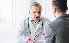 A doctor in a white coat and stethoscope gesticulates at he talks to a patient with his back to the viewer. 