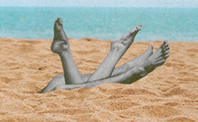 Two pairs of legs are coming out of the sand on a beach with the ocean in the background.