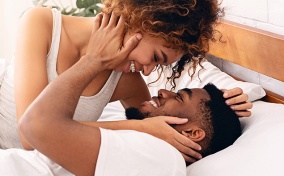A woman leans over a man in bed and they hold each other's faces, smiling.