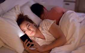 A woman lays in bed looking at social media while her male partner sleeps.