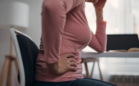 A woman in a pink shirt holds the side of her stomach with one hand and her head with the other.