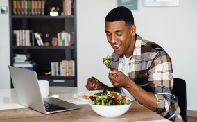 A man sits at his laptop eating a salad.