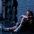 A bearded, shirtless man sits on the floor of a gym with his back against the wall. 