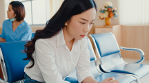 A woman sits in a medical waiting room looking down, concerned.