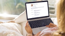 woman sits on white bed and looks at her resume on laptop