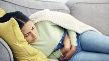 A woman lays on the sofa with a blanket over her as she holds her stomach in pain.