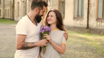 man and woman embrace in courtyard as they walk, he hands her a bouquet of purple flowers