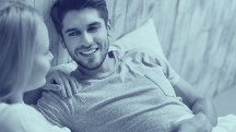 A new couple sits in bed smiling and talking to each other.