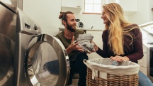 A man takes laundry out of the dryer and hands it to his female partner with a smile.