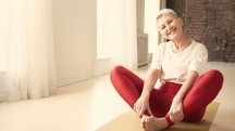 A menopausal woman in red pants and a white top is smiling and sitting on a yoga mat with her feet together.