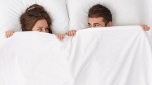 A man and woman lay beside each other in bed and hold the bedsheet up over their smiling faces.