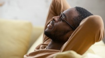 A black man leans back onto a sofa with his arms up behind his head.