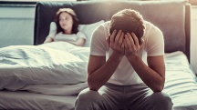 A man sits on the end of a bed with his head in his hands as a woman lays back on the opposite side.